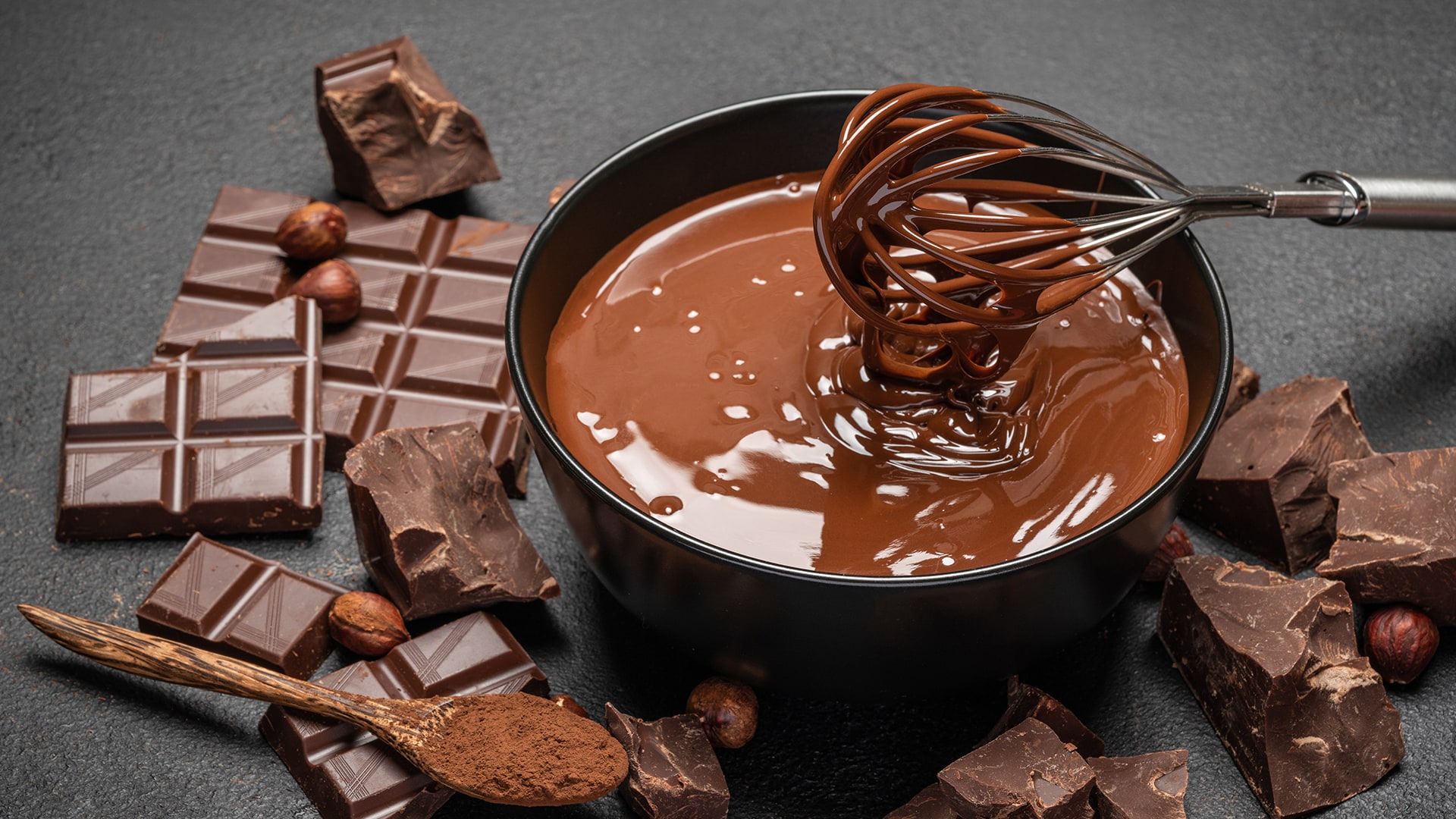 Schale mit flüssiger Schokolade und Schneebesen umringt von Schokoladenstücken
