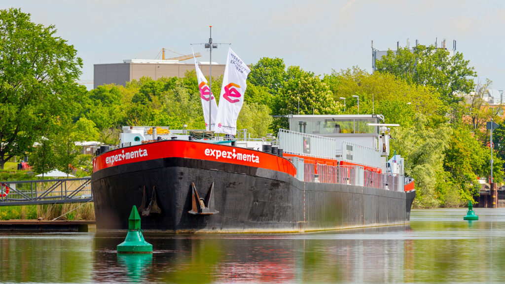 Das Schiff "ms-experimenta" liegt vor Anker, im Hintergrund grüne Vegetation.