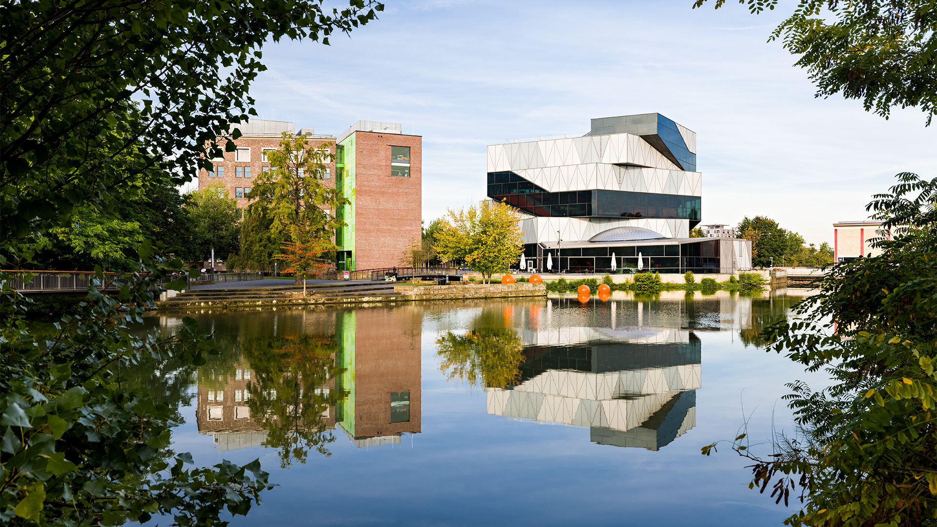 Die Gebäude der experimenta in Heilbronn spiegeln sich im ruhigen Wasser des nahegelegenen Sees. Umgeben von Bäumen und herbstlicher Natur.